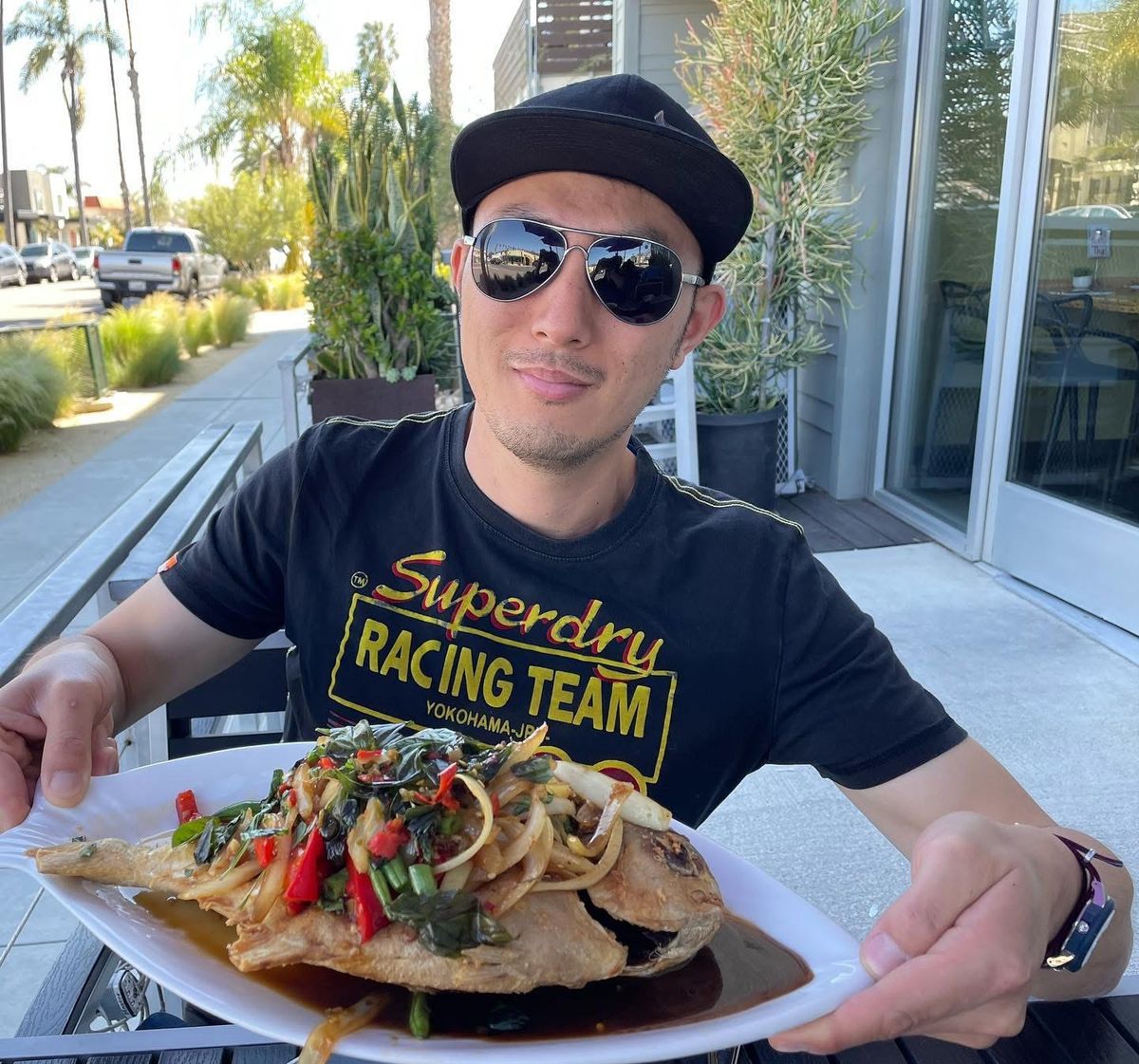 Roger on a sunny patio holding a whole fried fish topped with basil, chilies and onions